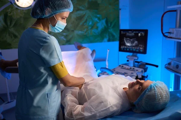 Uma médica se prepara para realizar a coleta de óvulos em uma paciente, que está deitada em uma sala de procedimentos, recebendo acompanhamento e cuidados da profissional.